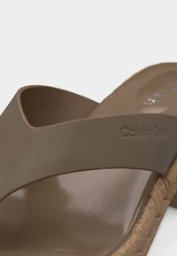 Κοντινό πλάνο μιας σαγιονάρας Calvin Klein σε αποχρώσεις του ταουπέ, με φαρδιά λουρίδα και λεπτομέρεια πλεγμένης σόλας.