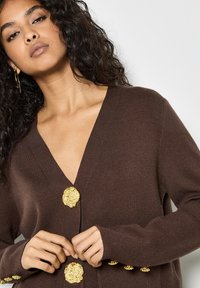 Cardigan marrone con design a V, grandi bottoni dorati a forma di fiore e polsini a coste. Il tessuto appare morbido e texturizzato.