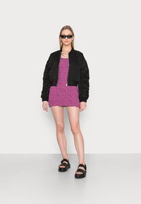 Jaqueta bomber preta curta, vestido mini texturizado roxo e sandálias pretas chunky. Modelo em pé sobre um fundo claro.