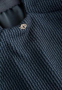 Marinblå stickad topp med texturerad, vågig tyg. Har en rund detalj i guld vid halsringningen. Mjuk, flexibel matrial.