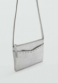 Silverfärgad fusk läderaxelväska med en strukturerad yta, dragkedjestängning och dekorativa nitar på framsidan. Justerbar rem ingår.