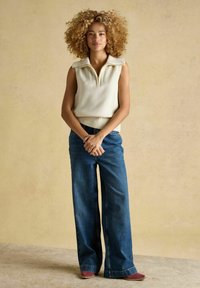 Hellbeige ärmelloser Pullover mit einem Reißverschlusskragen, kombiniert mit weitgeschnittenen blauen Jeans und roten Wildlederschuhen. Einfache Gestaltung und entspannte Passform.