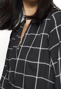 Černá blůza s bílým kostkovaným vzorem, s kulatým výstřihem a zdobením na knoflíky, vyrobená z měkké látky s hladkou texturou.