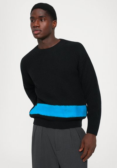 Schwarzer Strickpullover mit einem auffälligen blauen horizontalen Streifen in der Taille, runden Ausschnitt und langen Ärmeln über grauen Hosen.