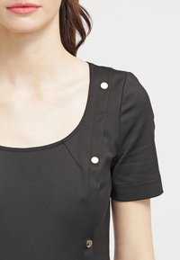 Sort kjole med rund halsudskæring, korte ærmer og sølvfarvede trykknapper på siden. Glat, mat tekstur med minimal syning.