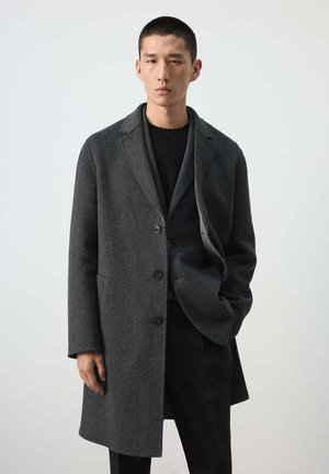 Un mannequin masculin se tient devant un fond uni, vêtu d’un pardessus gris foncé boutonné sur des vêtements noirs.