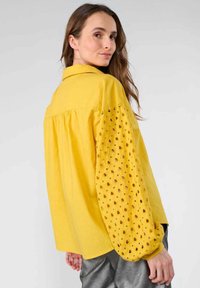 Camicia di cotone gialla con maniche lunghe e gonfie, caratterizzata da motivi a cuore ritagliati. Collo con bottoni e vestibilità ampia con dettaglio arricciato nella parte posteriore.