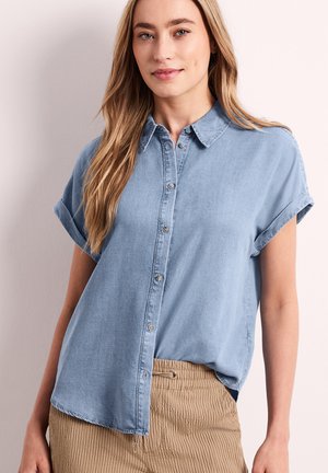 Ung kvinde med langt blondt hår iført en kortærmet blå skjorte og beige bukser med striber mod en lys baggrund.