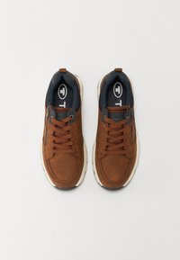 Ett par bruna avslappnade sneakers med snörning och sidodragkedjor, vadderad krage och vita sulor, visade uppifrån på en vit bakgrund.