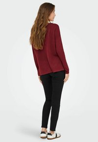 Maglione bordeaux a maniche lunghe con vestibilità morbida, spacchi laterali e polsini a coste, abbinato a jeans skinny neri e sneaker bianche.