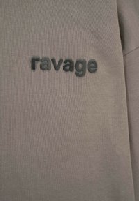 Brązowy materiał z wyhaftowanym słowem "ravage" w czarnych literach, wyśrodkowanym na powierzchni.