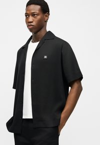 Ung mand iført en kortærmet sort skjorte med knapper over en hvid t-shirt, stående med den ene hånd i lommen mod en ensfarvet baggrund.