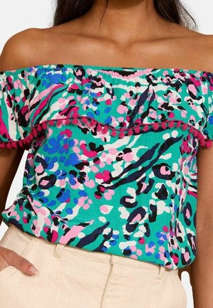 Femme portant un haut décolleté avec un motif floral abstrait teal, rose, blanc et noir, et un pantalon taille haute beige, la main dans la poche.