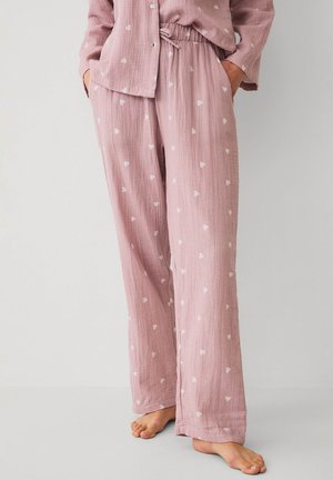 Weit geschnittene rosa Pyjamahose mit weißen Herzmustern, getragen mit passendem Oberteil, Hände in den Taschen, barfuß auf einem hellen Boden stehend.