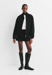 Žena u crnom oversized saku, crnoj mini suknji, bijelom crop topu i crnim čizmama do koljena stoji naslonjena na bijelu pozadinu.