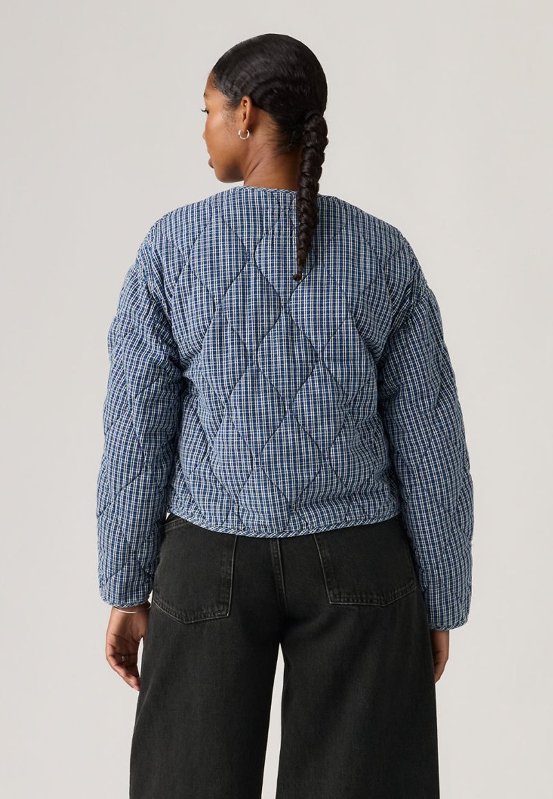 Femme avec une longue tresse portant une veste matelassée à carreaux bleue et un jean large noir, debout de dos sur un fond uni.