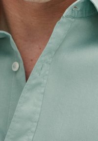 Jasnozielona bawełniana koszula z plisą zapinaną na guziki i kołnierzem. Tkanina ma gładką fakturę z kontrastowymi przeszyciami i guzikami.