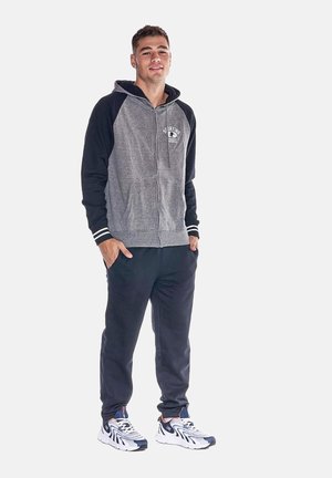 Jeune homme debout les mains dans les poches, portant un sweat à capuche gris et noir, un jogging foncé et des chaussures de course blanches et bleu marine.