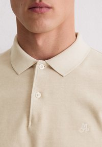 Beige Poloshirt mit weicher Textur, klassischem Kragen, drei Knöpfen und einem dezenten, in Weiß gestickten Logo auf der Brust.