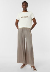 Kurzärmliges cremefarbenes T-Shirt mit "BREEZE" in goldener Schrift, kombiniert mit hellbraunen, weit geschnittenen Hosen und weißen Riemchensandaletten, vor einem neutralen Hintergrund.