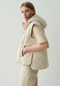 Beige gesteppte Pufferweste mit Kapuze, seitlichen Taschen und glatter Textur, kombiniert mit einem hellen Pullover und cremefarbenen Hosen.