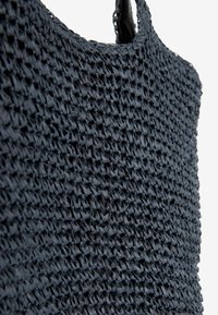 Navy modrá tkaná slaměná taška s texturovaným povrchem, vybavená kulatou rukojetí a prostorným otevřeným designem pro nošení věcí.