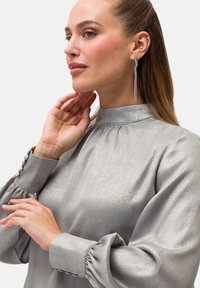 Blusa metallica argentea con colletto alto e maniche a sbuffo, tessuto texturizzato e dettagli con bottoni ai polsini; abbinata a lunghi orecchini scintillanti.