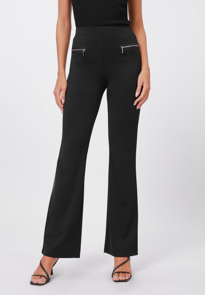 Zwarte flare broek met een hoge taille, voorzien van twee voorzakken met ritssluiting. Gemaakt van een gladde, rekbare stof. Gecombineerd met zwarte sandalen met bandjes.