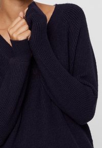 Merino-villaneule, jossa on V-pääntie, leveät hihat ja ribbitekstuuri; ominaisuuksia pitkät hihansuut, jotka peittävät osan käsistä.