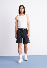 Black cargo shorts with two front pockets, adjustable waistband, paired with a white tank top and white sneakers, standing on a blue floor.