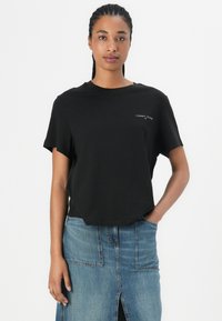 Svart t-shirt med kort ärm tillverkad av bomull, med rundad halsringning och en diskret logotyp på vänster sida. Matchad med en jeanskjol.