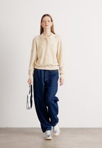 Maglione beige con colletto e decorazioni floreali abbinato a jeans a palazzo blu scuro e sneakers argentate, mentre tiene in mano una borsa a specchio.