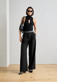 Top negro sin mangas con un agujero en el pecho, pantalones negros de cintura alta, cinturón grueso y bolso negro. Modelo con gafas de sol de gran tamaño.