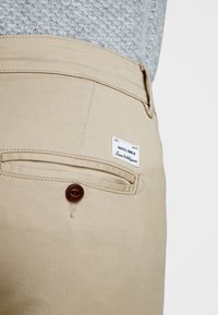 Pantalones de algodón beige con un bolsillo trasero que cuenta con un cierre de botón marrón y una etiqueta blanca con la marca "Jack & Jones". Tejido texturizado.