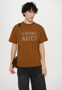 Brown cotton t-shirt with short sleeves, featuring a glittery "J'ADORE ARIES" print. Paired with dark jeans and a brown bag.