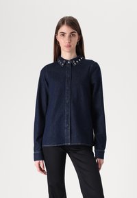 Camicia in denim di un blu scuro con chiusura a bottoni frontale, colletto decorato con borchie in metallo e maniche lunghe. Presenta cuciture a contrasto.