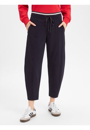 Persoon draagt zwarte broek met aantrekkoord, rood shirt met lange mouwen en witte sneakers met zwarte strepen, staat met handen in de zakken.