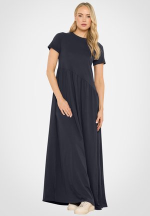 Mujer rubia con vestido largo azul marino de manga corta y zapatillas blancas, de pie frente a un fondo claro y uniforme.