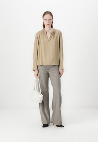 Haut beige à manches longues avec un décolleté en V profond, associé à un pantalon large gris. Le mannequin tient un petit sac blanc et porte des chaussures noires à bouts pointus.