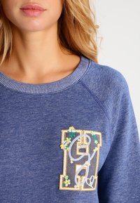 Sudadera azul con cuello acanalado y acentos florales bordados en un parche con letra, con un acabado de tela texturizada.