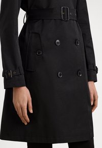 Black trench coat with double-breasted buttons, waist belt, and side pockets. Features smooth texture and adjustable cuff straps.