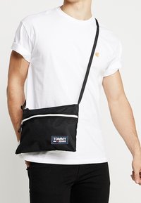Borsa a spalla in rete nera con zip bianca, dotata di un applicazione a patch con il logo "TOMMY JEANS" blu e rosso, tracolla regolabile, forma rettangolare.