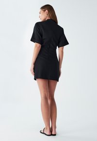 Črna kratek rokav obleka s prilegajočim krojem in zadrgo na zadnji strani. Gladko blago, dolžina do sredine stegen in stranski razpoki za gibljivost.