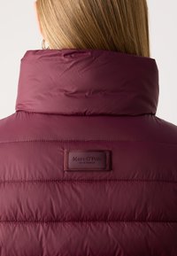 Sirge blonde juustega naine kannab kõrge kraega, fikseeritud burgundi värvi sulejope, mille seljal on Marc O'Polo logo plaaster.