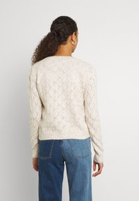 Kobieta z kręconymi włosami w niskim kucyku, nosząca srebrne kolczyki w kształcie kół, beżowy sweter w romby oraz dżinsy w dwóch odcieniach niebieskiego, odwrócona plecami.