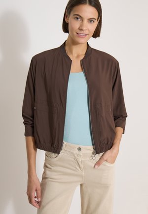 Vrouw met een bruine jas met rits over een lichtblauw topje en beige broek, staand met één hand in de zak tegen een lichte achtergrond.