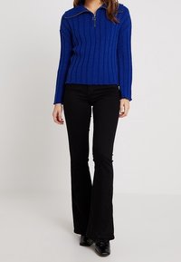 Blå ribbad zip-up-tröja med hög krage, tillsammans med svarta utsvängda byxor och glansiga svarta skor.