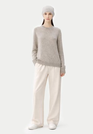 Frau steht, trägt einen hellgrauen Strickpullover, passende Mütze, weite cremefarbene Hose und weiße Sneakers vor einfarbigem Hintergrund.
