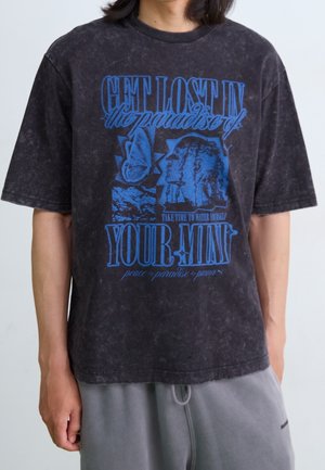 Schwarz gewaschenes T-Shirt mit blauem grafischem Text „VERLIERE DICH im Paradies DEINES GEISTES“, Schmetterlings- und Profilillustrationen, getragen zu grauer Hose.