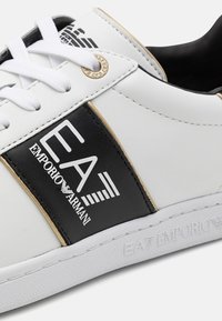 Białe skórzane sneakersy z czarnym panelem z logo. Złote akcenty wzdłuż szwów oraz białe sznurowadła dodają kontrastu do projektu.